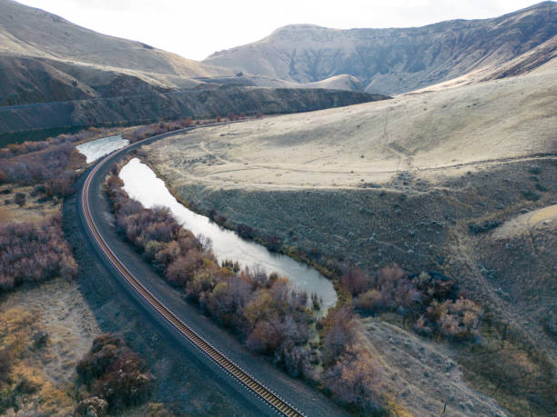 Beautiful train tracks winding through golden shrubby hills alongside a blue river with treed vegetation and sun shining.