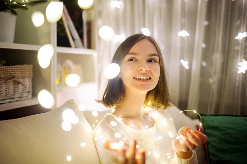 Beautiful teenage girl playing with Christmas lights in a cozy living room on Christmas eve. Celebrating Xmas at home. Winter evening with family and kids.