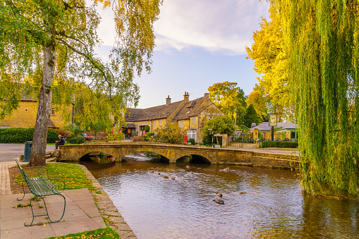 Bourton-on-the-Water, UK - October 17, 2022: Sunset scene of typical houses, the river Windrush, locals and visitors, in the village Bourton-on-the-Water, the Cotswolds region, England, UK
