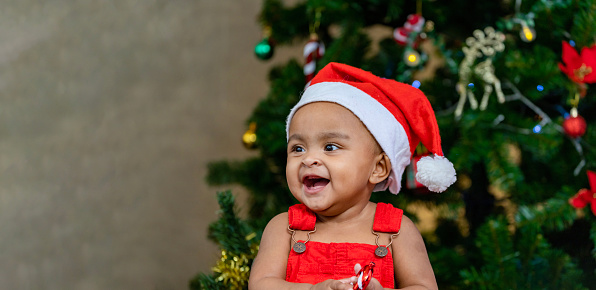 African American toddler baby is happily smiling while while dressing in christmas clothes and santa hat with christmas tree on the back for season celebration