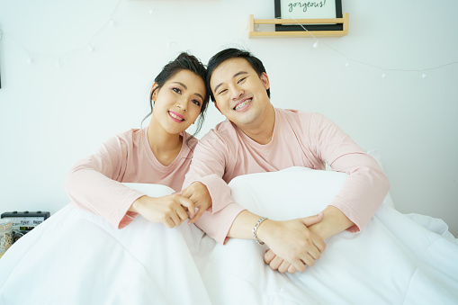 Happy and romantic Asian couple's portrait in bedroom with natural light from window, concept of relationship between husband and wife and being a family.