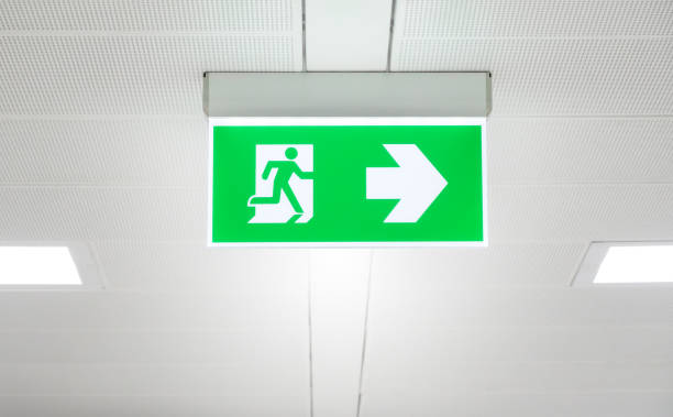 istock Lighted glowing green emergency exit signs in an office hallway with arrows pointing the way out of the building. 1448270800