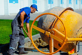 Uniformed construction worker works on construction site on summer day. Elderly bricklayer shovels cement and sand into concrete mixer. Contractor for repair of buildings and structures.