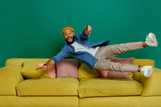 Excited young man using remote control while jumping on the couch against green background stock photo