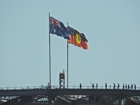 The Australian and Australian Aboriginal flags fly on the Sydney Harbour Bridge on an afternoon in summer, with silhouettes of people climbing the bridge.   This image was taken from Mrs Macquarie's Chair.