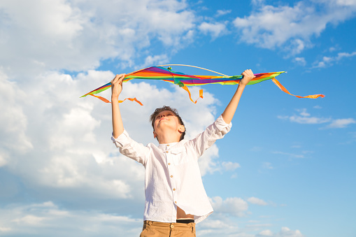 A 9-year-old boy with a bright colorful kite stands high mountain against a background of beautiful clouds and looks into the distance.