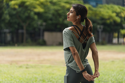 Active young Asian sportswoman listening to music with headphones, stretching arms and shoulders outdoor