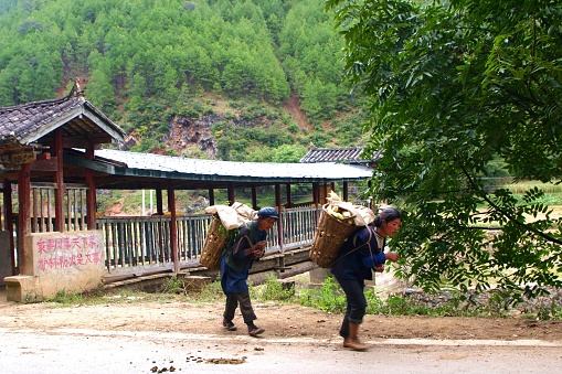 There was corn in the basket. Village people carry heavy loads on their backs and walk over covered Bridge and country road. tribes(ethnic) are unknown. Photo in 10/20/2007, Weixi County, Yunnan