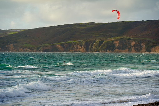 Kiteboarding with red kite on the water. Water sportsman with twintip board between waves in front of mountain landscape. France, Normandy, Manche, Vauville.