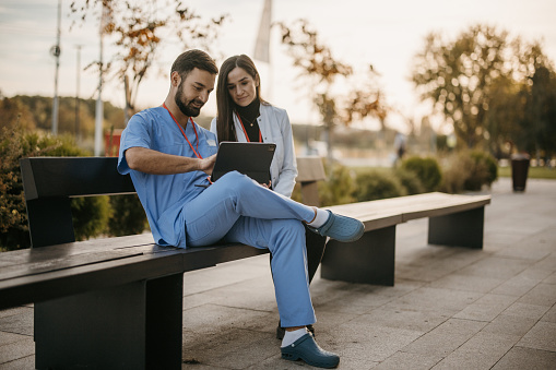 Two male and female doctors sitting on a bench in front of hospital and using digital tablet