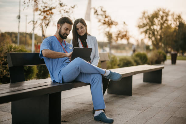 Male and female doctors sitting on a bench in front of hospital and using digital tablet stock photo