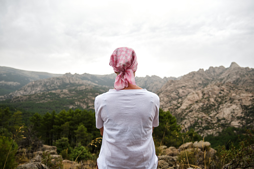 Woman with cancer sitting in a natural space. She wears a pink scarf on her head