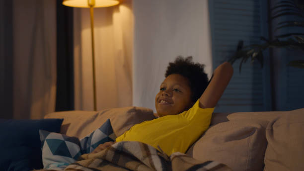 African-American kid sitting alone on sofa holding remote control and watching tv African-American kid sitting alone on sofa holding remote control and watching tv. Portrait of African-American preteen boy relaxing on couch and changing channels on tv in evening at home tremoctopus gelatus stock pictures, royalty-free photos & images