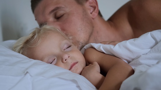 The father kisses his sleeping daughter on the shoulder, gently looking at her, then kisses her on the head again, covers his daughter with a blanket and calmly falls asleep next to her in bed.