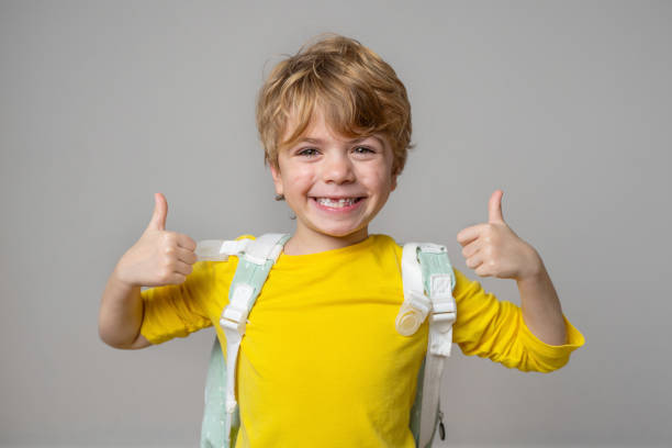 primary school smiling boy girl over grey background with backpack showing thumbs up. copy space for advertising blank concept. back to school. childhood, education, products for children - elementary school building imagens e fotografias de stock