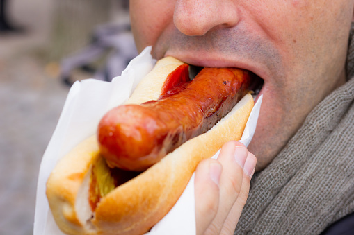Close up of mouth of man eating hot dog with ketchup and mustard. Classic German sausage, fast food concepts