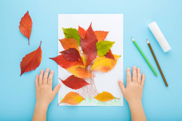 istock Little child hands creating tree shape from colorful leaves on white paper. Light blue table background. Pastel color. Making autumn decorations. Point of view shot. Closeup. Top down view. 1448321742
