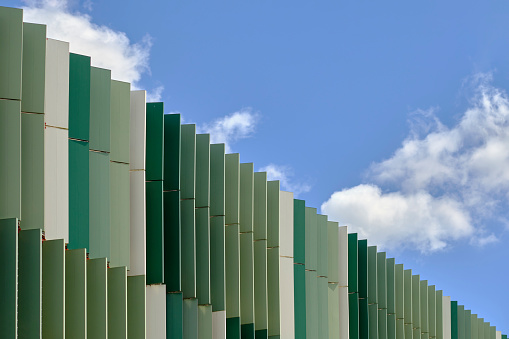 Metal panels in shades of greens. Blue sky with white fluffy clouds. Frame divided diagonally between the building's facade and the sky