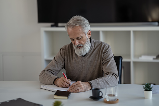 A gray-haired bearded man in casual clothes sits at a desk in front of a large TV in an office and looks over notes in a black book.