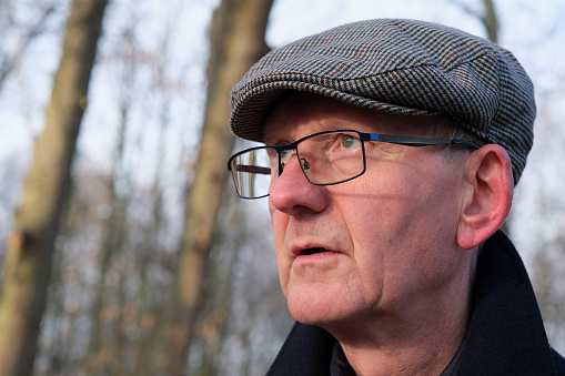 Portrait of mature, elderly or mature adult Caucasian male in woodland wearing a flat cap. Cold winter or autumn day outdoors. Essex, United Kingdom