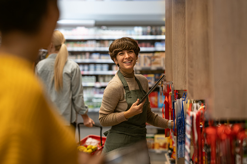A smiling saleswoman in an apron is talking to a customer with a friendly smile at the shelves with goods.