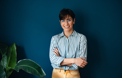 Portrait of a happy young businesswoman with short hair, wearing glasses. She is leaning against a wall with her arms crossed and looking at the camera.