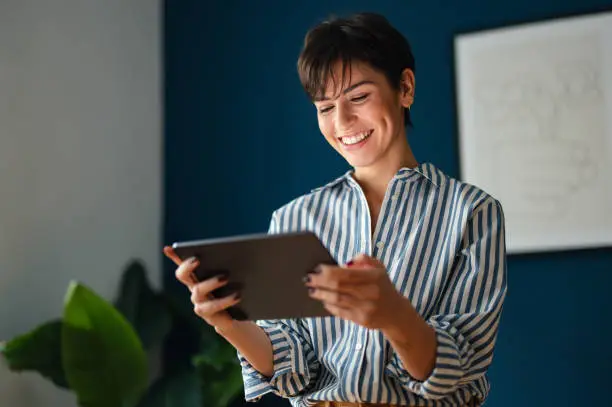 Portrait of a beautiful modern businesswoman leaning against the desk in her office. She is doing some work on her digital tablet while looking down at it and smiling.