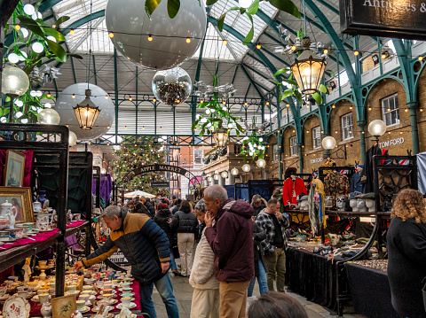 People browsing the antique stalls in the Apple Market in London’s Covent Garden Market. Christmas baubles and oversized mistletoe are hanging from the domed ceiling.