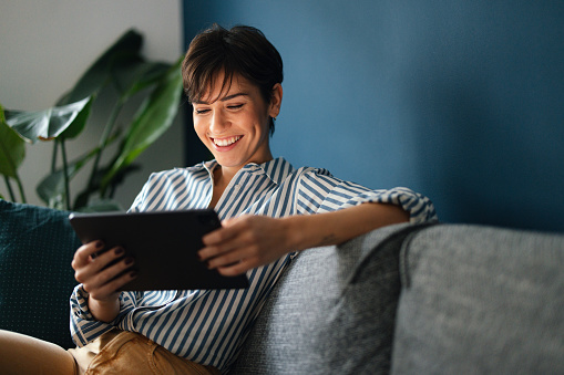 Close up shot of a beautiful smiling young woman with short hair sitting comfortably on the sofa at home. She is looking down and using her digital tablet.