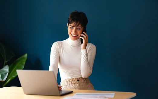 Close up shot of a beautiful smiling businesswoman with short hair, sitting at the work desk and talking with someone on her mobile phone while looking down at her laptop computer.