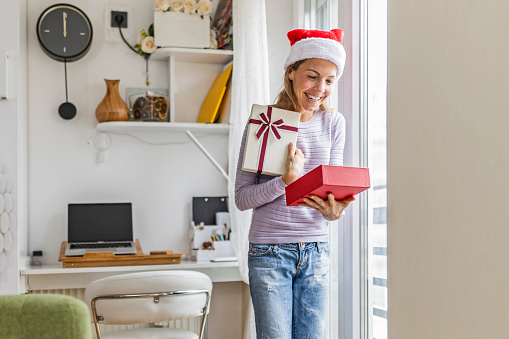 Young woman in Christmas mood standing next to a window in living room holding gift box