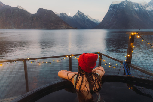 Vista trasera de una mujer con sombrero rojo relajándose en la bañera de hidromasaje con vista panorámica del fiordo de invierno en Noruega photo