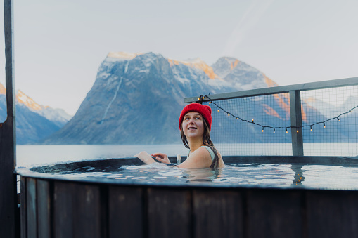 Vista lateral de una mujer feliz con sombrero rojo relajándose en la bañera de hidromasaje con vista panorámica del fiordo de invierno en Noruega photo