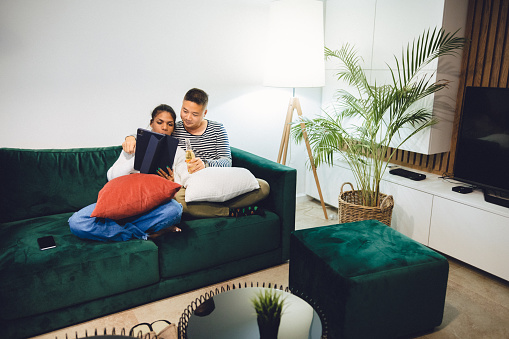 Multiracial heterosexual couple enjoying media at home while sitting on a sofa and using a digital tablet.