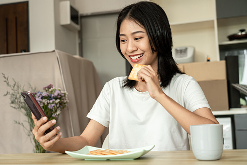 Asian woman eats crackers for breakfast and uses her mobile phone to check the morning news before going to work, Working people's urgent food, Urgent activity after waking up.