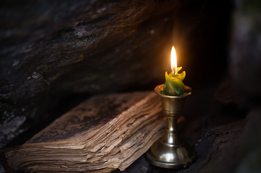 Magic theme. Burning candle and old book in stone dark niche in the forest, close up, selective focus. Foreboding, November, autumn, witchcraft, mystery, victorian style, gothic concept