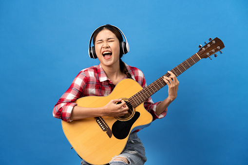 Young asian woman wearing headphone and gingham red shirt with braid hairstyle while playing guitar and singing rock song with happiness isolated over light blue background.