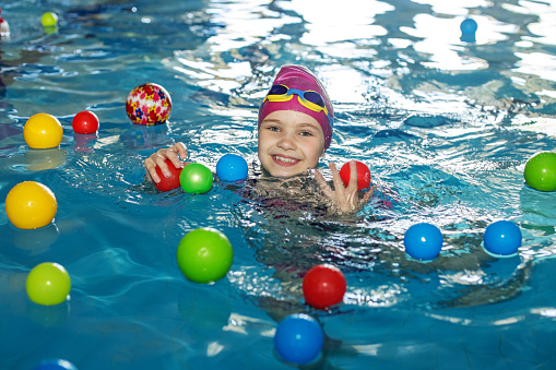 Happy child schoolgirl is studying at swimming lessons in pool. Swimming cap and goggles. Lots of colorful balls. Concept of healthy lifestyle and sports.