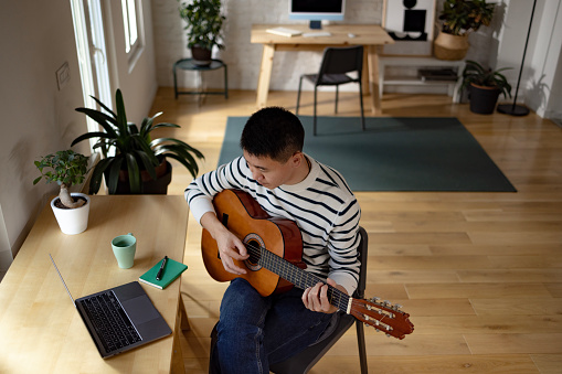 Above view of young Asian man teaching guitar lesson during a video call over a computer at home.