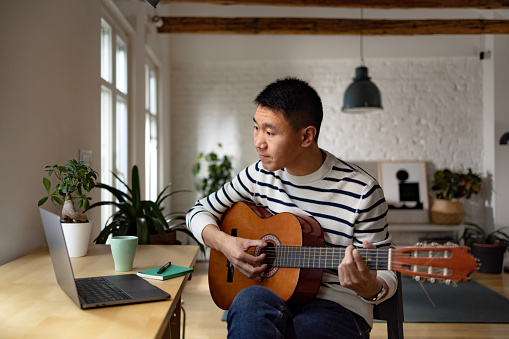 Asian man having an online guitar lesson at home.