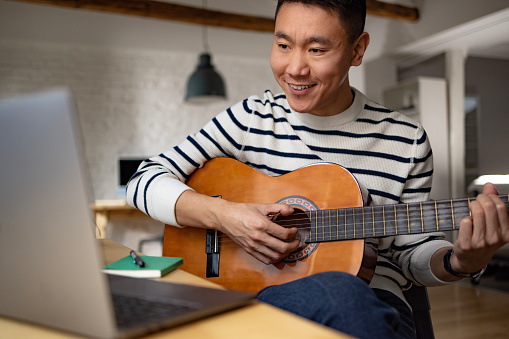 Happy Asian man having an online guitar lesson over a computer at home.