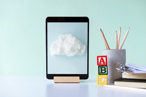 Front view of a white desk with a digital tablet, pen holder with pens, notepad,paperclip, and toy blocks with the letters A,B,C in front of blue wall. There is an image of a cloud  on device screen. Representing e-learning.