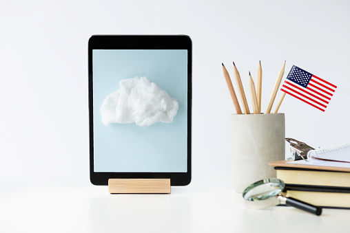 Front view of a white desk with a digital tablet, pen holder with pens and an American flag , notepad,paperclip, and magnify glass. There is an image of a cloud  on device screen. Representing e-learning in the USA.