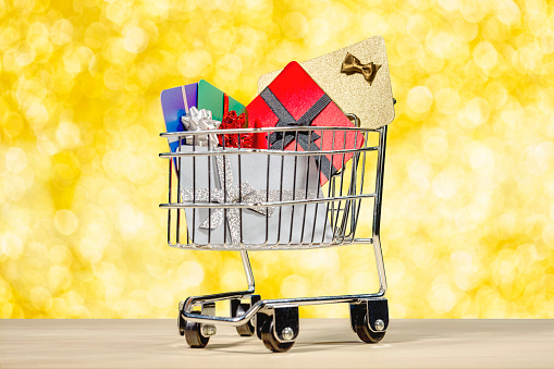 A miniature shopping cart filled with wrapped gift cards against a gold colored bokeh background.