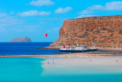 Crete, Greece - October 4, 2021: Scenic top view of Balos lagoon with people, red kitesurf and big ship below.