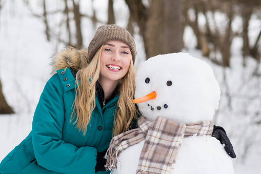 Beautiful blonde young woman wearing a winter coat is smiling for a photo with the snowman she built in the winter forest. He has a carrot nose, button eyes and a plaid scarf.