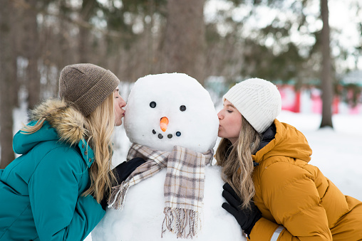 Two beautiful young women are kissing the cheeks of the snowman they built in the snowy public park. They are wearing warm winter coats and hats. The snowman has buttons for his eyes and mouth, carrot nose and a scarf around his neck.