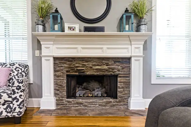 Close up of a white wood fireplace in a new home with windows and a chair in a living room.