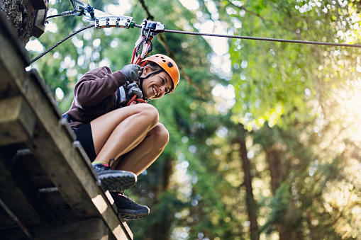 Teenage girl enjoying high ropes course in adventure park. 
The girl is wearing safety harness and a helmet. The girl is getting ready to zip lining using tyrolean traverse from one platform to another.
Sunny summer day.
Canon R5