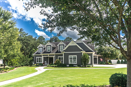A large gray craftsman new construction house with a landscaped yard and leading pathway sidewalk on a sunny day with blue skies and clouds.